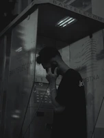A person stands inside a dark and dimly lit phone booth, holding a phone receiver to their ear. The atmosphere is moody with shadows dominating the scene. The booth has the name 'BAKTELECOM' on it and contains a keypad and other typical phone booth features.