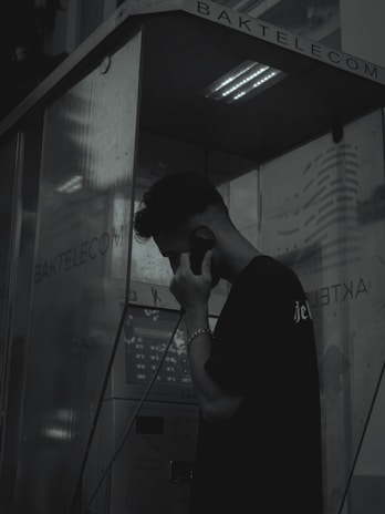 A person stands inside a dark and dimly lit phone booth, holding a phone receiver to their ear. The atmosphere is moody with shadows dominating the scene. The booth has the name 'BAKTELECOM' on it and contains a keypad and other typical phone booth features.
