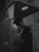 A person stands inside a dark and dimly lit phone booth, holding a phone receiver to their ear. The atmosphere is moody with shadows dominating the scene. The booth has the name 'BAKTELECOM' on it and contains a keypad and other typical phone booth features.