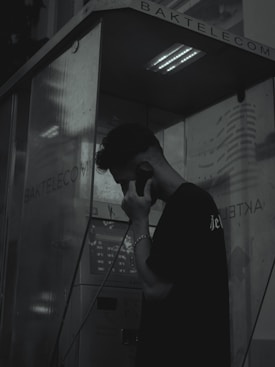 A person stands inside a dark and dimly lit phone booth, holding a phone receiver to their ear. The atmosphere is moody with shadows dominating the scene. The booth has the name 'BAKTELECOM' on it and contains a keypad and other typical phone booth features.