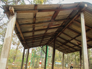 A rustic, open wooden structure with a corrugated metal roof supported by wooden pillars. The surroundings consist of a forest with numerous trees. In the background, there are a few painted logs and a metal ladder structure. The roof appears weathered and aged.