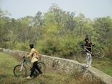 Two people are outdoors in a natural setting with lush green trees and grass. One person is walking a bicycle along a narrow stone pathway while the other carries their bicycle over the path. The area is surrounded by a forest, giving a sense of adventure and exploration.