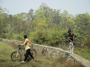Two people are outdoors in a natural setting with lush green trees and grass. One person is walking a bicycle along a narrow stone pathway while the other carries their bicycle over the path. The area is surrounded by a forest, giving a sense of adventure and exploration.