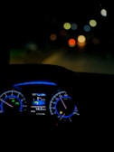 A dimly lit car interior focusing on the dashboard with illuminated speedometer and tachometer. The digital display shows average fuel economy and temperature information. The background is blurred, with colorful circular bokeh lights visible through the windshield.