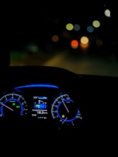 A dimly lit car interior focusing on the dashboard with illuminated speedometer and tachometer. The digital display shows average fuel economy and temperature information. The background is blurred, with colorful circular bokeh lights visible through the windshield.