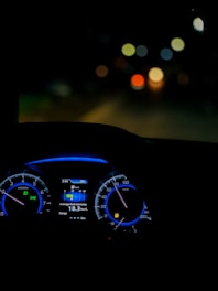 A dimly lit car interior focusing on the dashboard with illuminated speedometer and tachometer. The digital display shows average fuel economy and temperature information. The background is blurred, with colorful circular bokeh lights visible through the windshield.
