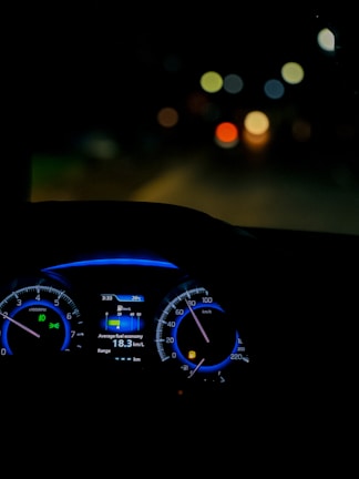 A dimly lit car interior focusing on the dashboard with illuminated speedometer and tachometer. The digital display shows average fuel economy and temperature information. The background is blurred, with colorful circular bokeh lights visible through the windshield.