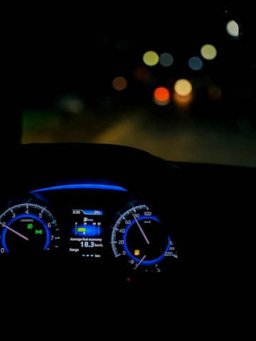 A dimly lit car interior focusing on the dashboard with illuminated speedometer and tachometer. The digital display shows average fuel economy and temperature information. The background is blurred, with colorful circular bokeh lights visible through the windshield.