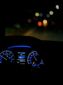 A dimly lit car interior focusing on the dashboard with illuminated speedometer and tachometer. The digital display shows average fuel economy and temperature information. The background is blurred, with colorful circular bokeh lights visible through the windshield.