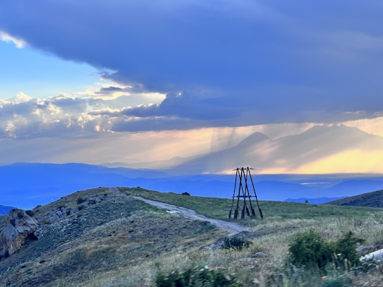 A serene landscape featuring rolling hills and grassy terrain, under a dramatic sky with clouds and rays of sunlight breaking through. In the background, silhouetted mountains create a misty, atmospheric effect. A wooden structure stands near a dirt path, adding a rustic element to the scene.