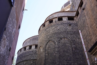 The image features the side of an ancient stone building with dark brickwork and arched architectural designs. The perspective is from below, looking upwards at the towering circular structures with a clear blue sky in the background. A modern building with worn plaster walls is partially visible on the left.