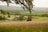 A scenic countryside landscape featuring farmland with a few buildings. The foreground includes tilled soil with some patches of grass, and a tree branch partially frames the top of the image. The midground has green fields and several farm structures, along with a red brick house. The background features rolling hills and a series of green and forested areas under a cloudy sky.