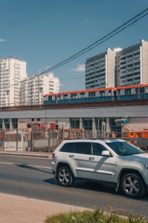 A busy street scene with a modern, white SUV driving along the road. Behind the car, a train with a red and blue design is traveling on an elevated track. In the background, there are several tall apartment buildings with white facades. The sky is clear and blue, suggesting a sunny day.