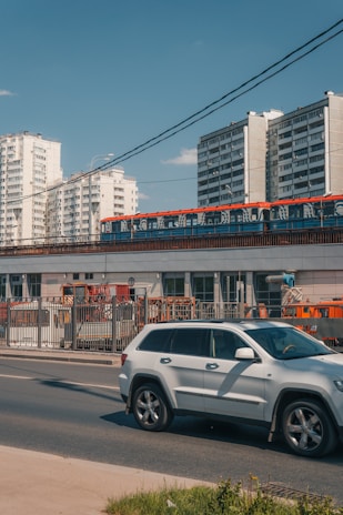 A busy street scene with a modern, white SUV driving along the road. Behind the car, a train with a red and blue design is traveling on an elevated track. In the background, there are several tall apartment buildings with white facades. The sky is clear and blue, suggesting a sunny day.