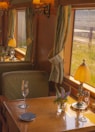 A vintage train carriage interior features wooden walls and green upholstered seats. Small tables have elegant turned lamps and clear vases with purple flowers. Champagne flutes rest on the table, and light filters through green curtains on large windows.
