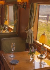A vintage train carriage interior features wooden walls and green upholstered seats. Small tables have elegant turned lamps and clear vases with purple flowers. Champagne flutes rest on the table, and light filters through green curtains on large windows.
