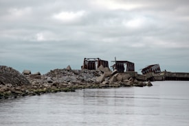 A coastal scene with remnants of a dilapidated structure made of rusted metal and concrete rubble. The area is surrounded by water, and the cloudy sky above creates a somber atmosphere. Rocks and debris line the shore, adding to the feeling of desolation.