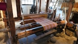 A traditional wooden loom is set up with colorful threads, positioned in a rustic interior space with stone floors and large windows providing natural light. Various weaving tools and materials are visible around the loom.