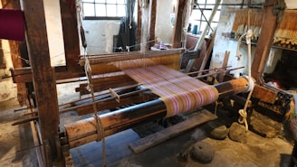 A traditional wooden loom is set up with colorful threads, positioned in a rustic interior space with stone floors and large windows providing natural light. Various weaving tools and materials are visible around the loom.