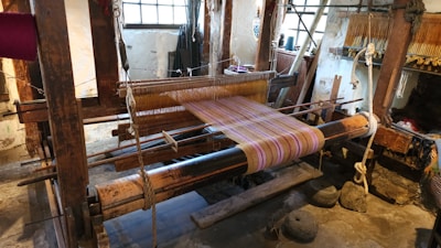 A traditional wooden loom is set up with colorful threads, positioned in a rustic interior space with stone floors and large windows providing natural light. Various weaving tools and materials are visible around the loom.
