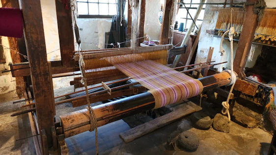 Close-up of skilled hands weaving green and cream threads on a traditional wooden handloom bathed in soft natural light.
