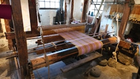 A traditional wooden loom is set up with colorful threads, positioned in a rustic interior space with stone floors and large windows providing natural light. Various weaving tools and materials are visible around the loom.
