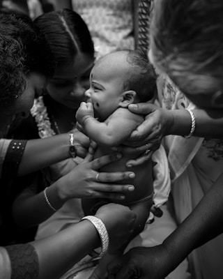 A group of women are gathered around a baby, holding it gently. The baby has a small mark on the cheek and appears to be sucking its thumb. The women's hands are adorned with bangles and rings, suggesting a cultural or family gathering.