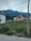 Two residential houses with red roofs are situated near a grassy field, backed by lush, green mountains under a cloudy sky. Power lines stretch across the scene, and a dirt path runs in the foreground.