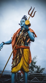 A large statue of a blue-skinned figure holding a trident. The figure wears an orange shawl and yellow garment, with detailed facial features and ornamentation. The background is a cloudy sky, and there are trees visible in the distance.