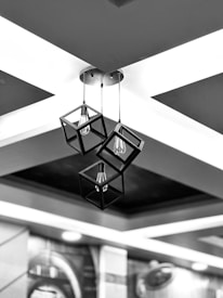 Three geometric pendant lights with a modern design hang from the ceiling. The lights are encased in cube-shaped frames, and each contains a bulb inside. The ceiling features a minimalistic cross pattern with contrasting black and white areas.