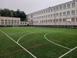 An outdoor sports field with artificial turf, marked with various lines for different sports, is set in front of a large, multi-story school building. The building features numerous white-framed windows and has a neutral color scheme. Surrounding the field are several basketball hoops and soccer goals. Trees and greenery are visible in the background.
