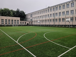 An outdoor sports field with artificial turf, marked with various lines for different sports, is set in front of a large, multi-story school building. The building features numerous white-framed windows and has a neutral color scheme. Surrounding the field are several basketball hoops and soccer goals. Trees and greenery are visible in the background.