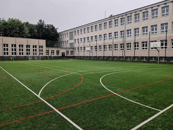 An outdoor sports field with artificial turf, marked with various lines for different sports, is set in front of a large, multi-story school building. The building features numerous white-framed windows and has a neutral color scheme. Surrounding the field are several basketball hoops and soccer goals. Trees and greenery are visible in the background.