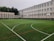 An outdoor sports field with artificial turf, marked with various lines for different sports, is set in front of a large, multi-story school building. The building features numerous white-framed windows and has a neutral color scheme. Surrounding the field are several basketball hoops and soccer goals. Trees and greenery are visible in the background.
