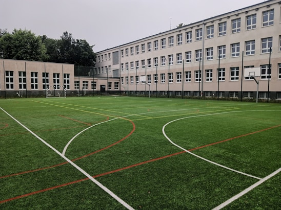 An outdoor sports field with artificial turf, marked with various lines for different sports, is set in front of a large, multi-story school building. The building features numerous white-framed windows and has a neutral color scheme. Surrounding the field are several basketball hoops and soccer goals. Trees and greenery are visible in the background.