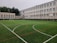 An outdoor sports field with artificial turf, marked with various lines for different sports, is set in front of a large, multi-story school building. The building features numerous white-framed windows and has a neutral color scheme. Surrounding the field are several basketball hoops and soccer goals. Trees and greenery are visible in the background.