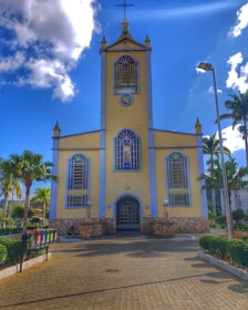 A yellow church with blue accents stands prominently under a vibrant blue sky. It features a central clock tower with a cross on top and arched windows. The surrounding area includes palm trees, neatly trimmed bushes, and a paved walkway leading to the entrance.