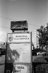 A black and white photograph of a public information board in an urban setting. The board displays the sign 'Municipiul Cluj-Napoca' and includes a map of the area. Above the sign, there is a digital display or billboard with visible numbers. Surrounding the board are streets, cars, and residential buildings, with some trees and wires visible in the background.