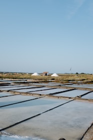 A landscape with large flat pools for evaporating seawater to extract salt. Piles of salt are visible in the distance near a small structure, with a vast, clear blue sky above. The scene is calm and expansive.