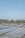A landscape with large flat pools for evaporating seawater to extract salt. Piles of salt are visible in the distance near a small structure, with a vast, clear blue sky above. The scene is calm and expansive.