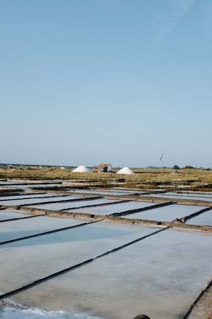 A landscape with large flat pools for evaporating seawater to extract salt. Piles of salt are visible in the distance near a small structure, with a vast, clear blue sky above. The scene is calm and expansive.