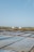 A landscape with large flat pools for evaporating seawater to extract salt. Piles of salt are visible in the distance near a small structure, with a vast, clear blue sky above. The scene is calm and expansive.