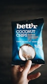 A hand holding a package of 'bett'r' coconut chips in salted caramel flavor. The packaging is predominantly blue with white text and an image of a coconut on it. The words 'baked', 'no added fat', and 'organic' are visible, along with 'guilt free'.