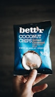 A hand holding a package of 'bett'r' coconut chips in salted caramel flavor. The packaging is predominantly blue with white text and an image of a coconut on it. The words 'baked', 'no added fat', and 'organic' are visible, along with 'guilt free'.
