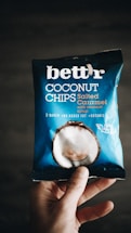 A hand holding a package of 'bett'r' coconut chips in salted caramel flavor. The packaging is predominantly blue with white text and an image of a coconut on it. The words 'baked', 'no added fat', and 'organic' are visible, along with 'guilt free'.