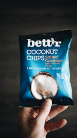 A hand holding a package of 'bett'r' coconut chips in salted caramel flavor. The packaging is predominantly blue with white text and an image of a coconut on it. The words 'baked', 'no added fat', and 'organic' are visible, along with 'guilt free'.