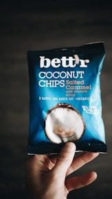 A hand holding a package of 'bett'r' coconut chips in salted caramel flavor. The packaging is predominantly blue with white text and an image of a coconut on it. The words 'baked', 'no added fat', and 'organic' are visible, along with 'guilt free'.