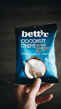 A hand holding a package of 'bett'r' coconut chips in salted caramel flavor. The packaging is predominantly blue with white text and an image of a coconut on it. The words 'baked', 'no added fat', and 'organic' are visible, along with 'guilt free'.