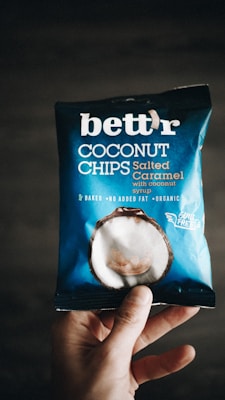 A hand holding a package of 'bett'r' coconut chips in salted caramel flavor. The packaging is predominantly blue with white text and an image of a coconut on it. The words 'baked', 'no added fat', and 'organic' are visible, along with 'guilt free'.