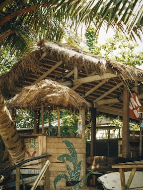 A rustic beach hut with thatched roof sits under the shade of palm trees. The structure is made of wood, featuring a simple counter and decorative elements like a painted surfboard and tropical motifs. A relaxed atmosphere permeates the scene, aided by the presence of surrounding greenery.